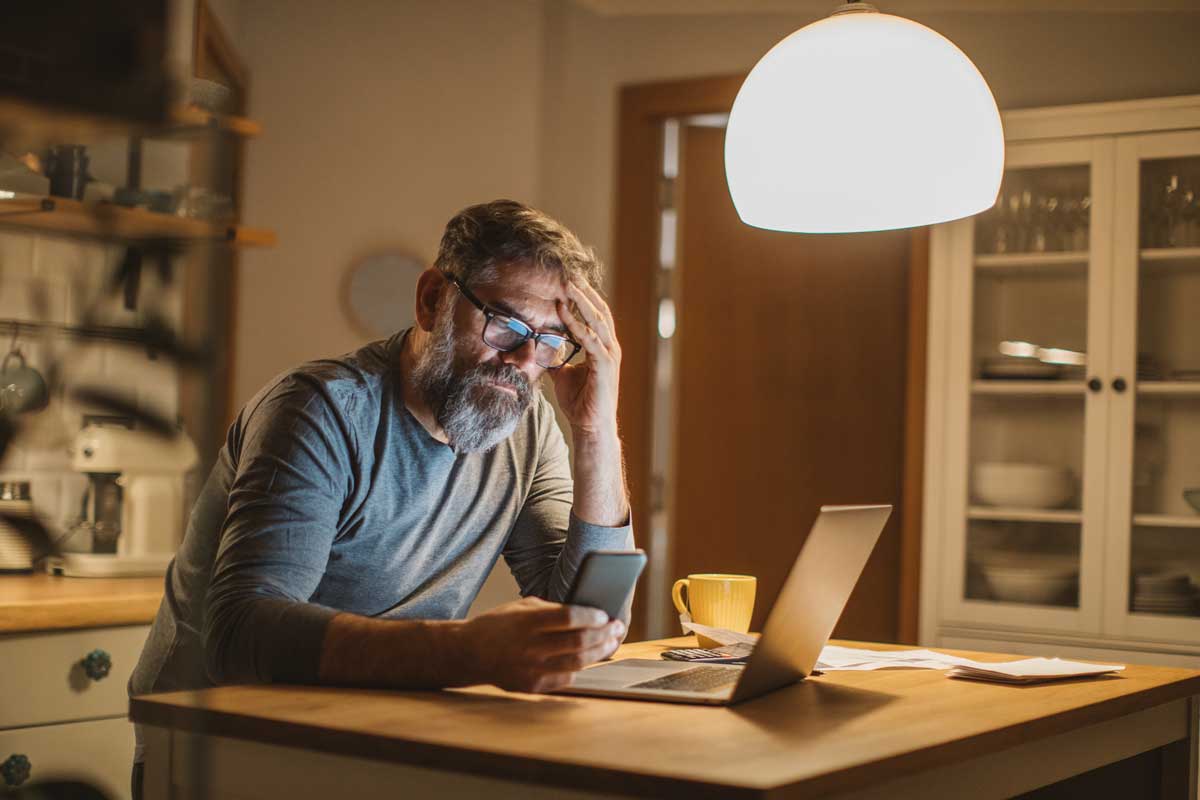 An upset man scrolls through his phone while sitting at the kitchen table in front of an open laptop.