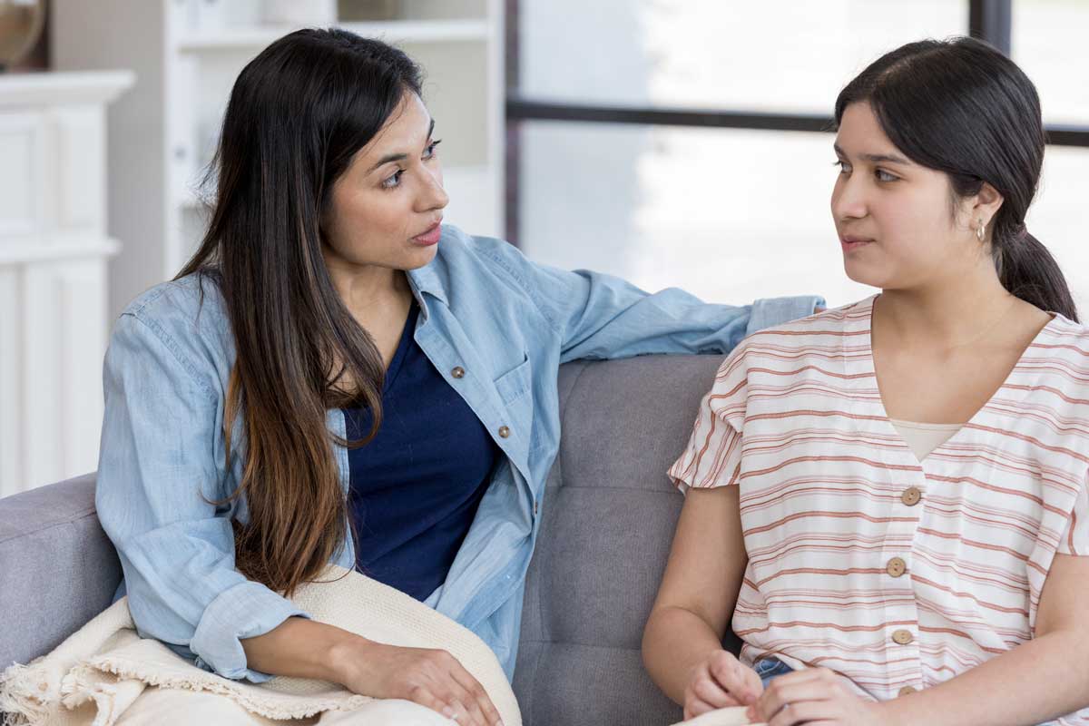 Mother and daughter have a serious conversation sitting on a couch in their home. 